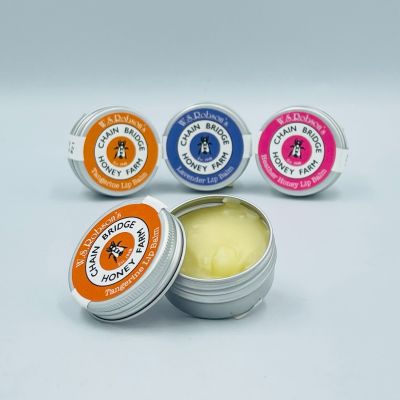 A row of three Chain Bridge honey and beeswax lip balms in a small tins with a branded stickers, also an open tin in front showing the balm inside, white background